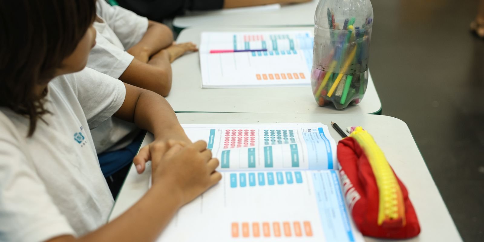 Estudantes de escola pública sentados em carteiras com material didático sobre a mesa