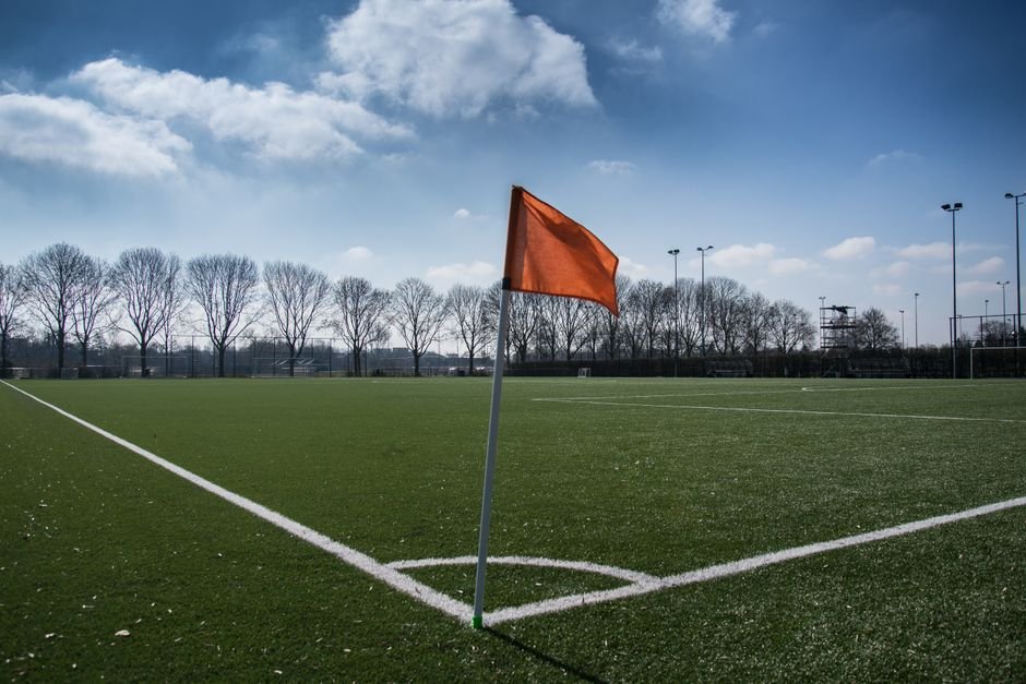 Campo de futebol gramado com bandeira de escanteio laranja em primeiro plano e traves ao fundo
