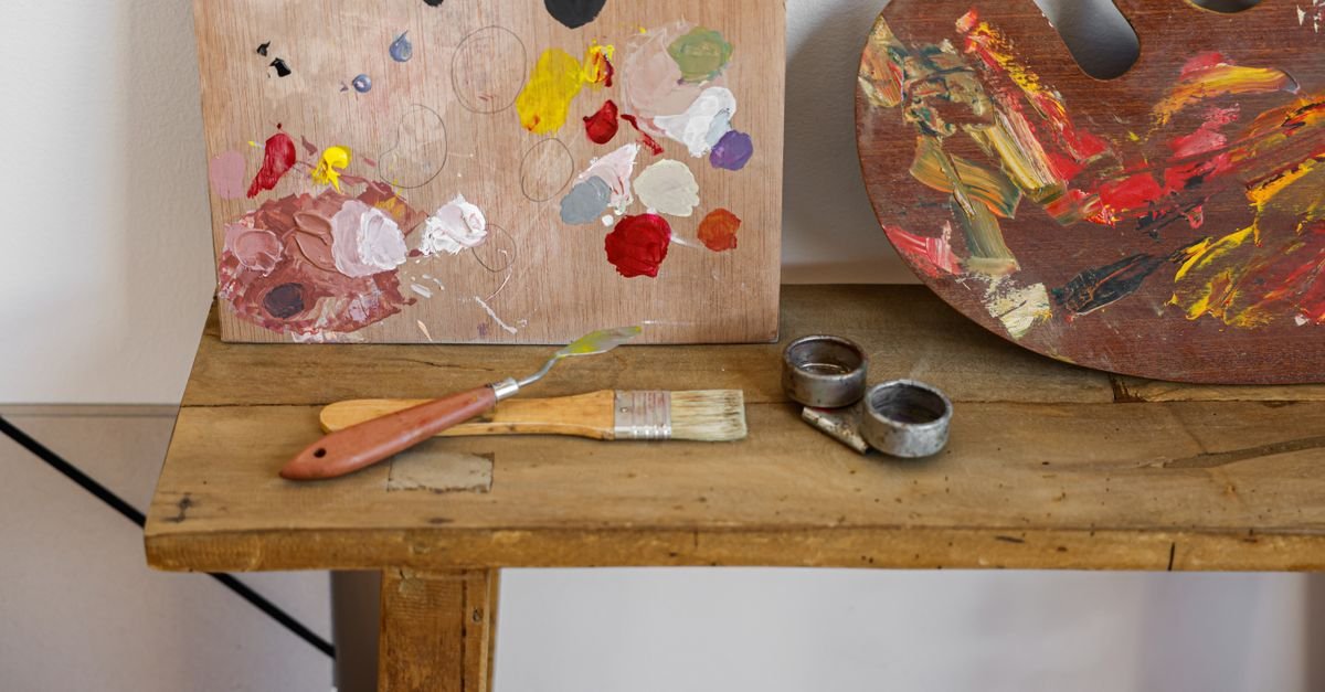 Paleta de pintura, pincel e espátula sobre bancada de madeira em ateliê