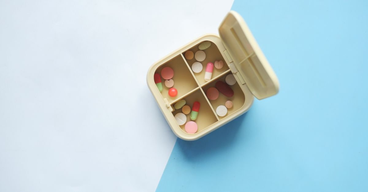 Caixa organizadora de medicamentos aberta, com comprimidos e cápsulas de cores variadas, vista de cima sobre fundo azul e branco