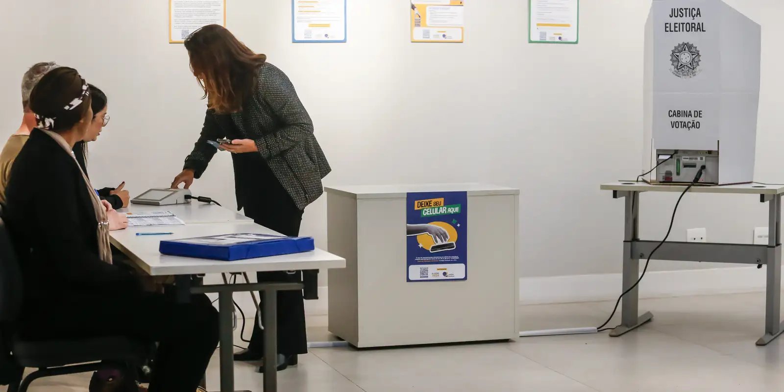 Mesa receptora de votos com cartaz 'Deixe seu celular aqui' ao lado da cabine de votação da Justiça Eleitoral