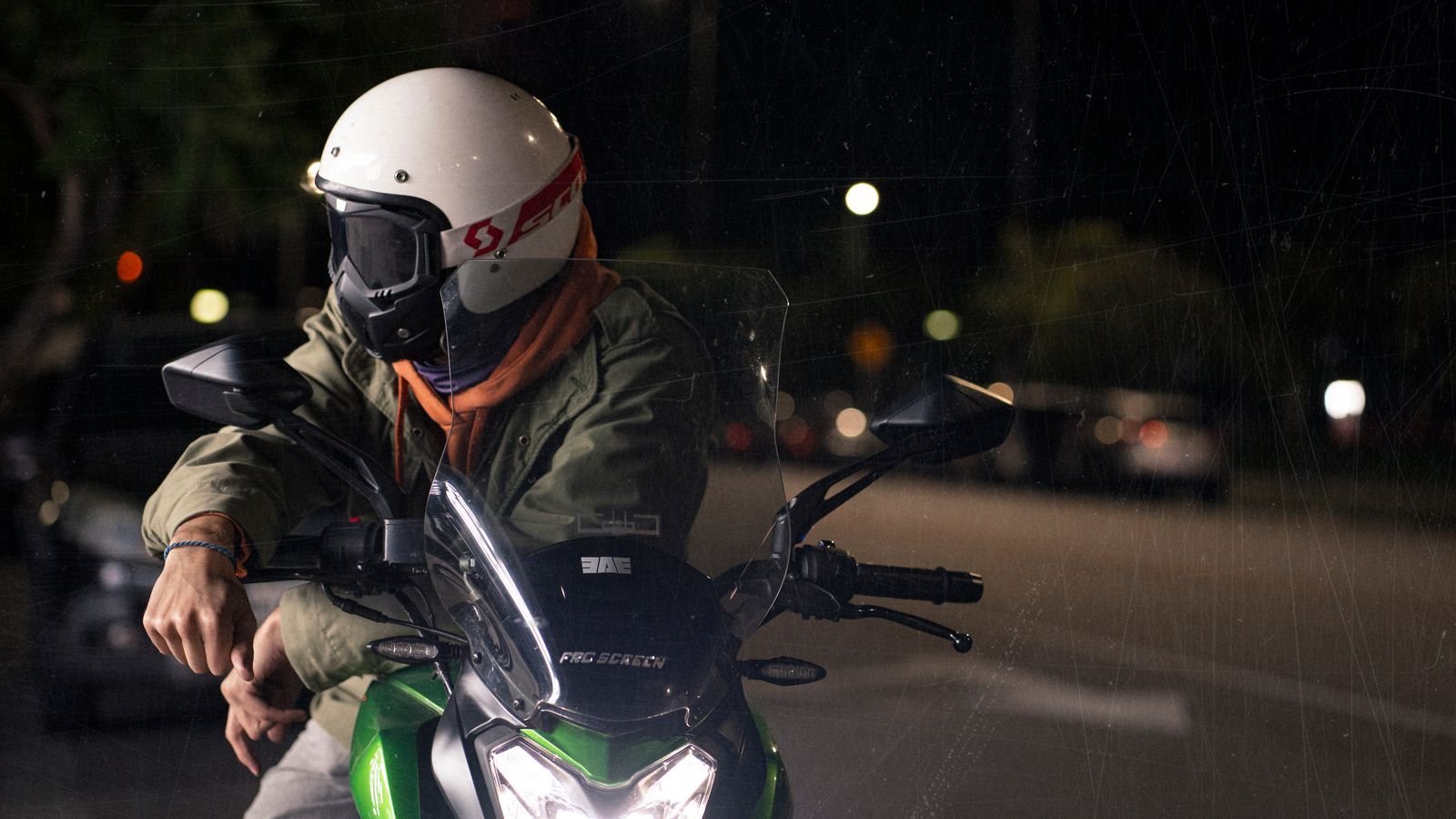 Motociclista de capacete branco parado em via urbana à noite, com faróis de carros ao fundo