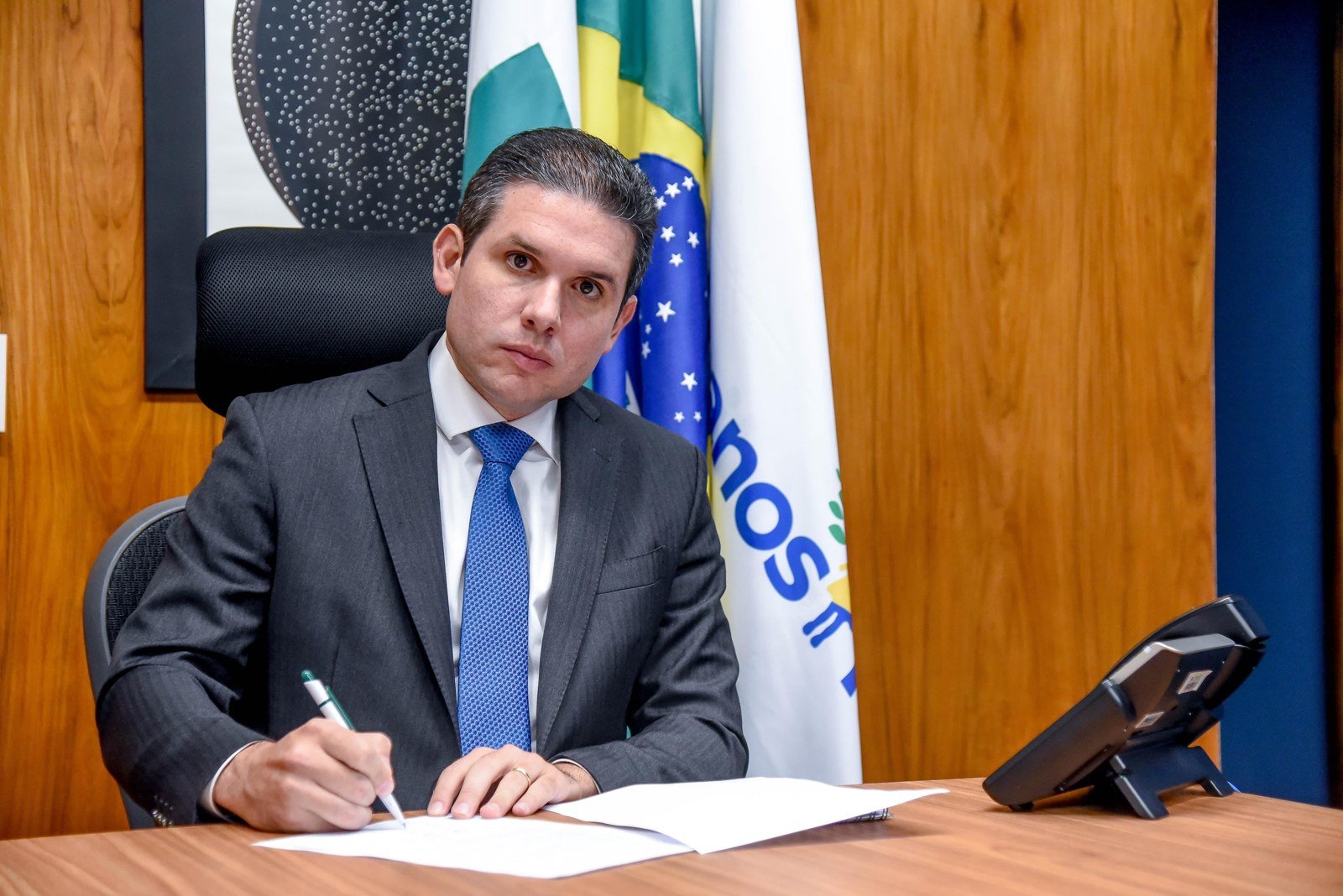 Hugo Motta assina documento em seu gabinete, com bandeiras ao fundo