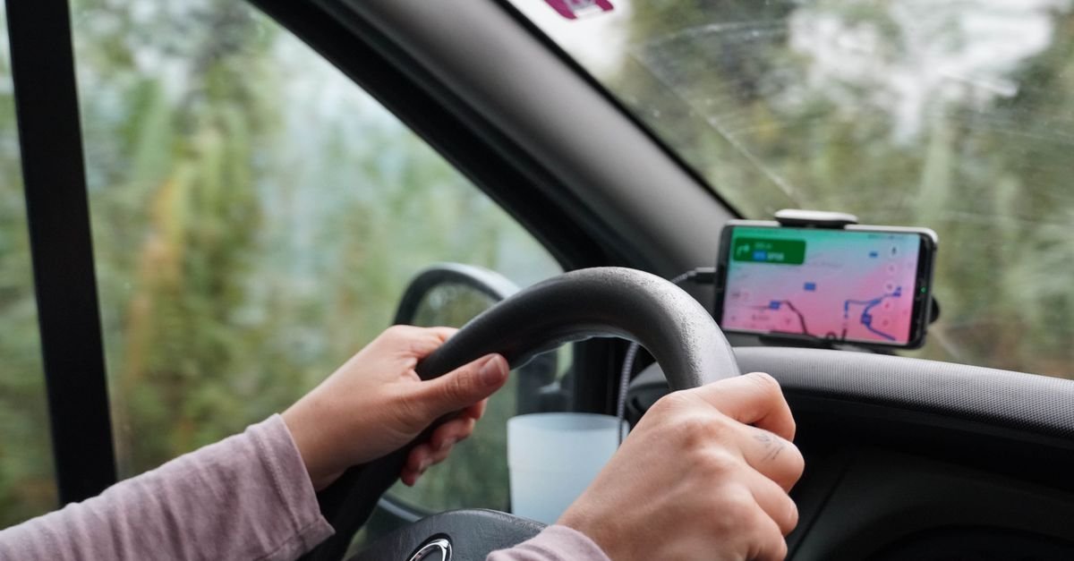Mãos ao volante de carro com celular em suporte exibindo aplicativo de navegação