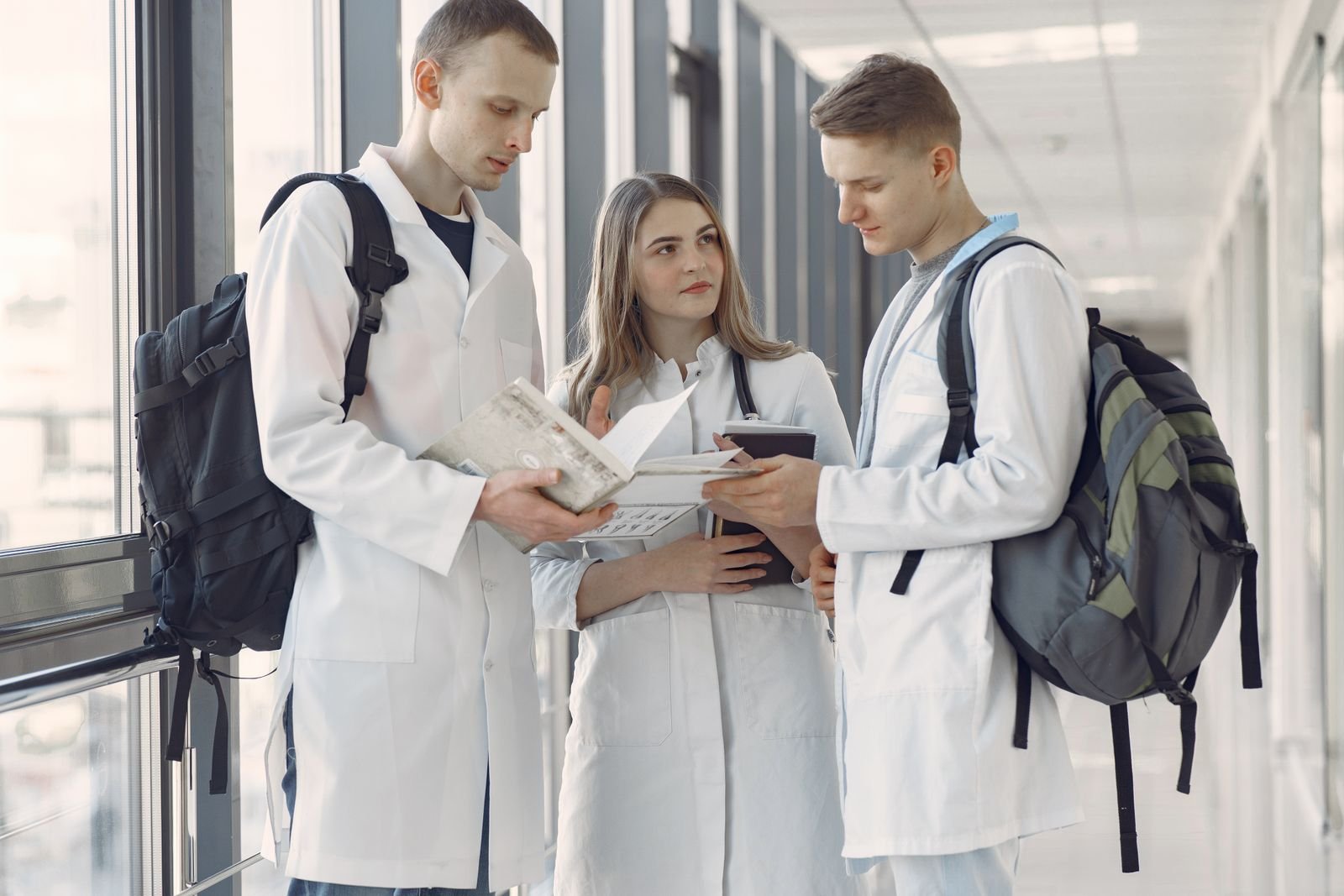 Três estudantes de jaleco branco, com mochilas e livros, conversam em um corredor (foto ilustrativa)