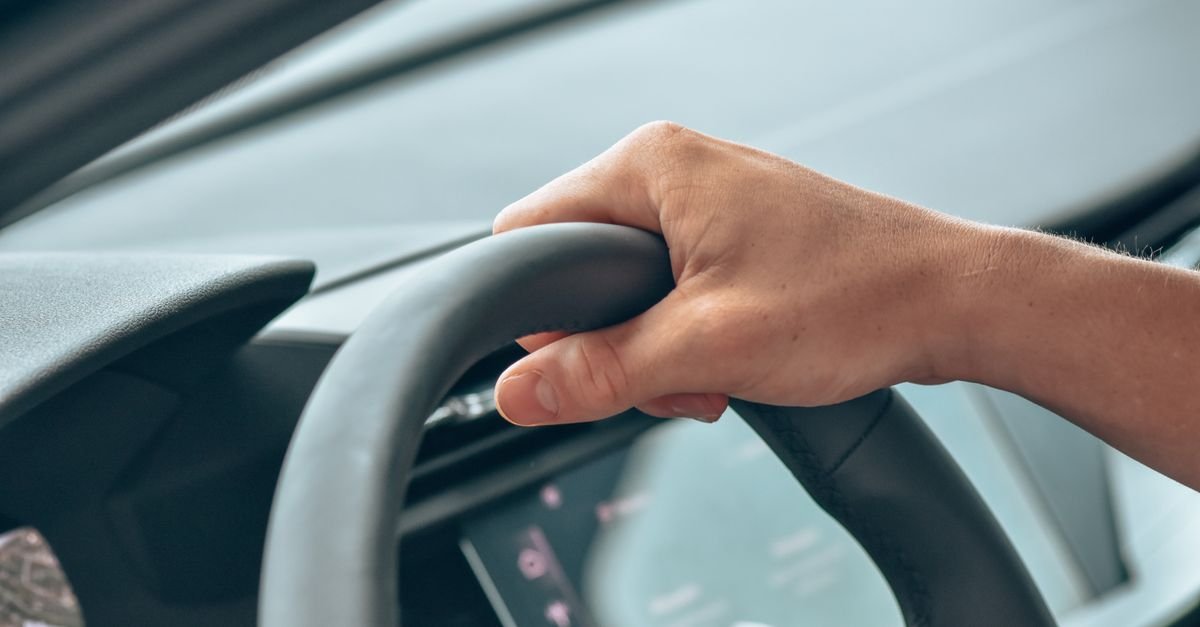Mão de motorista apoiada no volante de um carro, com o painel desfocado ao fundo