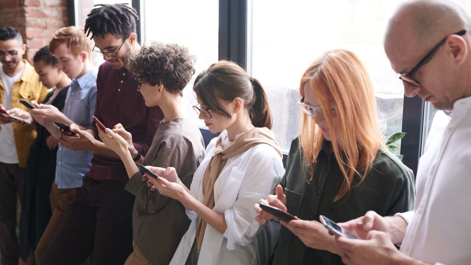Grupo de pessoas lado a lado, todas olhando para o celular, diante de uma janela