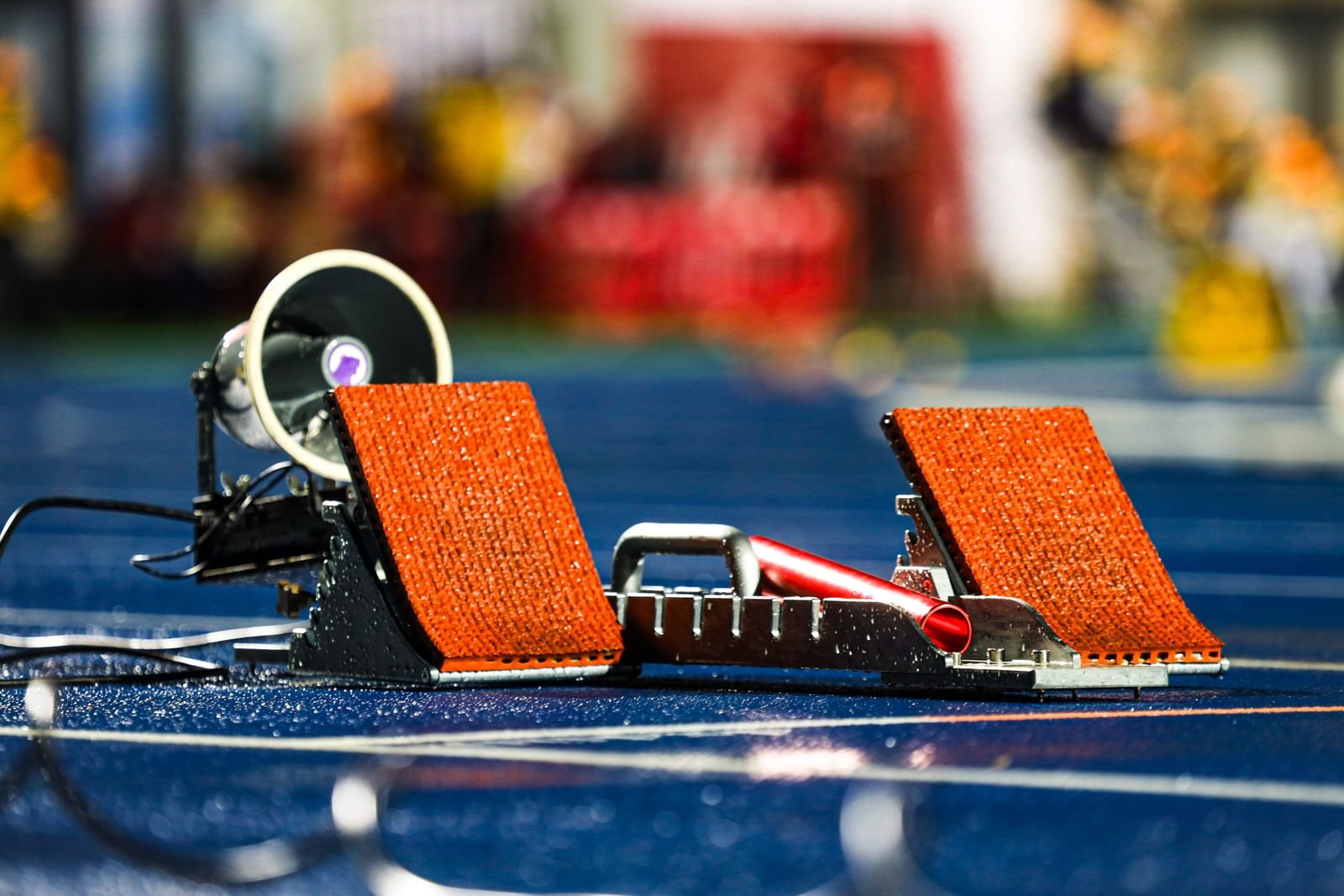 Blocos de largada laranja sobre pista de atletismo azul molhada, com megafone ao lado