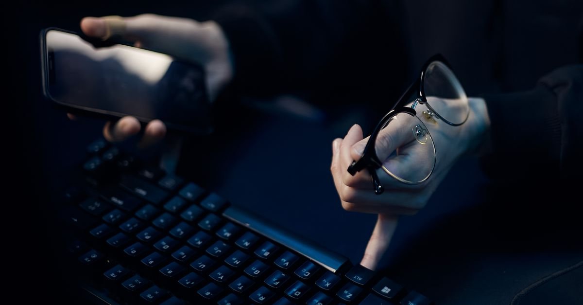 Mãos de uma pessoa segurando um celular ao lado de um teclado de computador em ambiente escuro