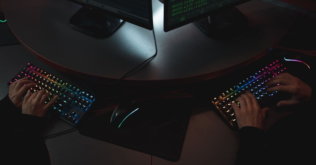 Duas pessoas digitam em teclados iluminados diante de telas com código