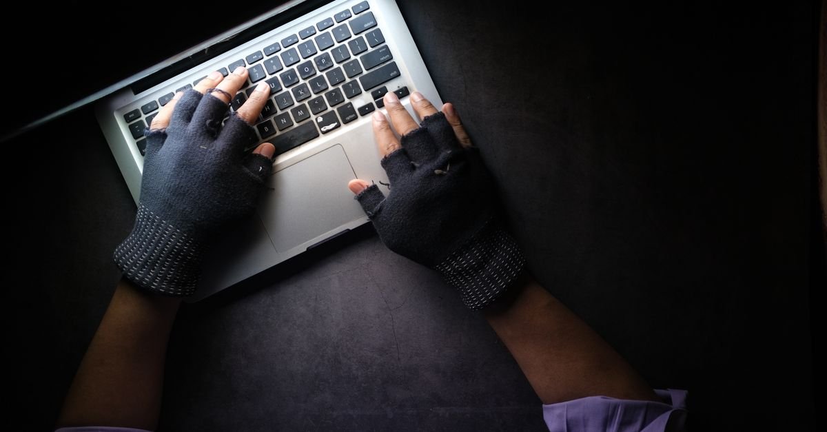 Mãos com luvas sem dedo digitando no teclado de um notebook em ambiente escuro