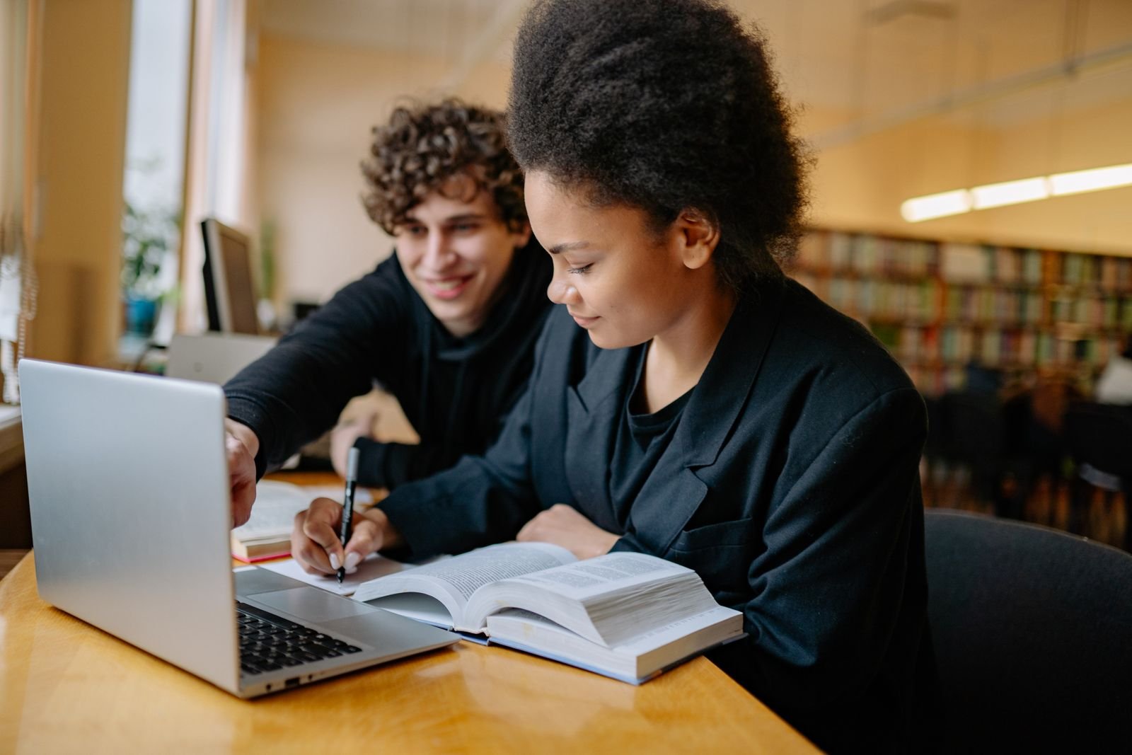 Dois estudantes universitários estudam juntos em uma biblioteca, com notebook e livro abertos