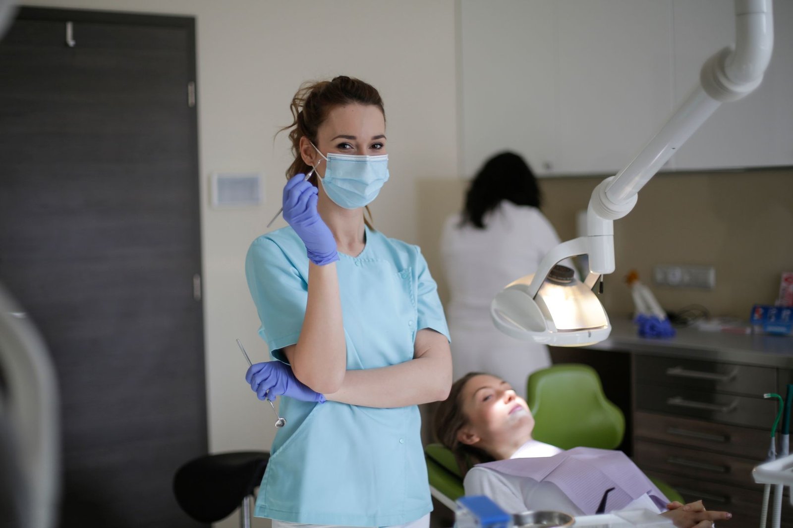 Profissional de odontologia com máscara, luvas e instrumentos em pé num consultório, com paciente na cadeira e colega ao fundo