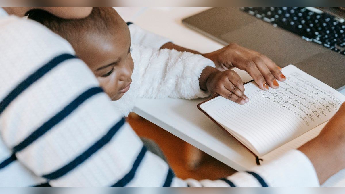 Mulher escreve em caderno com um bebê no colo, ao lado de um notebook sobre a mesa