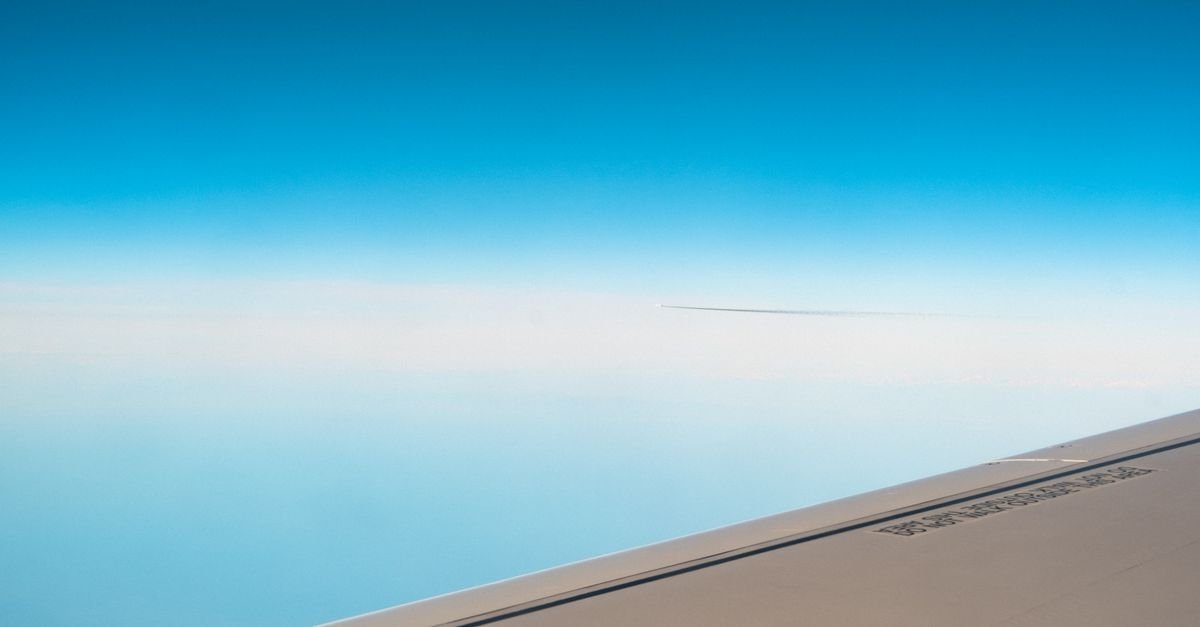 Asa de avião em voo vista pela janela, com céu azul e camada de nuvens ao fundo