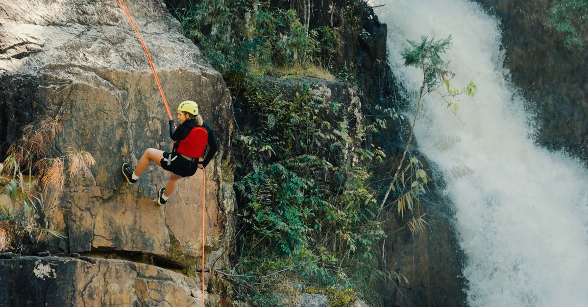 Pessoa de capacete desce de rapel por parede de rocha ao lado de cachoeira cercada de mata