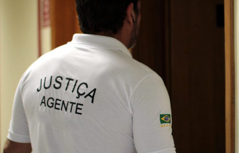 Agente da Justiça de costas, com a inscrição Justiça Agente na camisa, em corredor de fórum