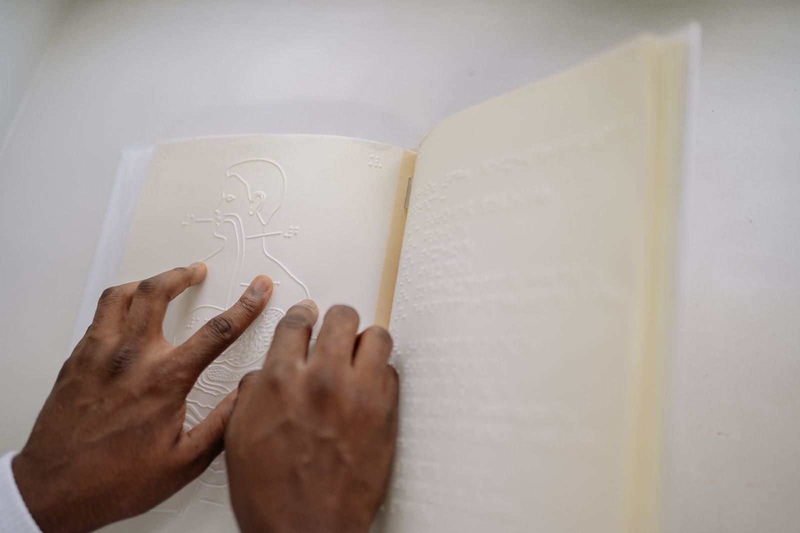 Mãos de uma pessoa leem com os dedos um livro em braille com ilustração em relevo; imagem ilustrativa