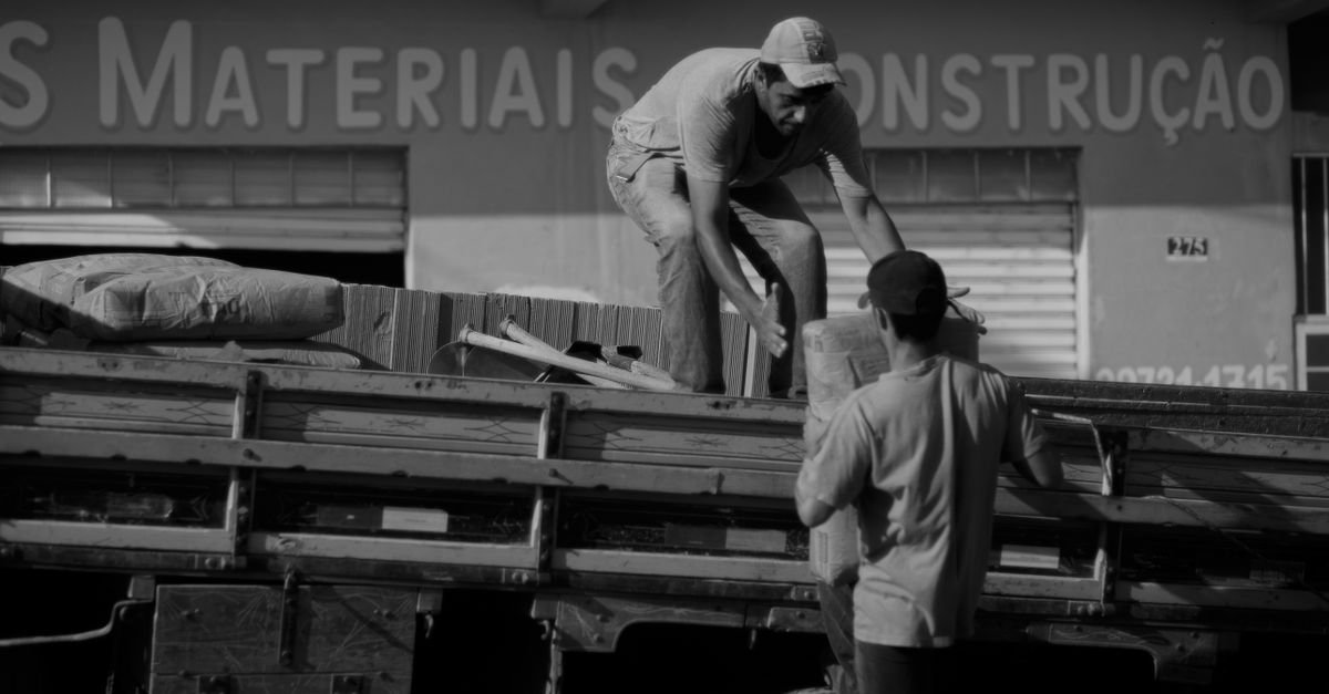 Dois trabalhadores descarregam material sobre a carroceria de um caminhão em frente a uma loja de materiais de construção