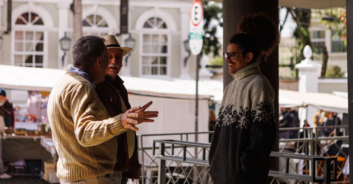 Tres pessoas idosas conversam em uma feira ao ar livre em dia de sol