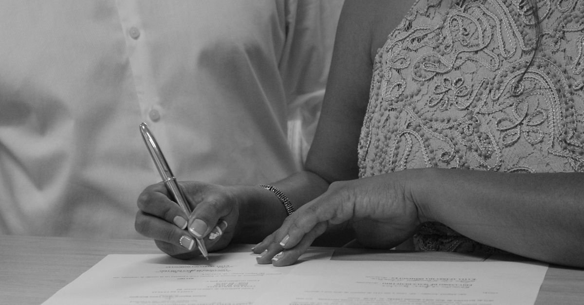 Mãos de duas pessoas assinando documento sobre mesa, em imagem em preto e branco