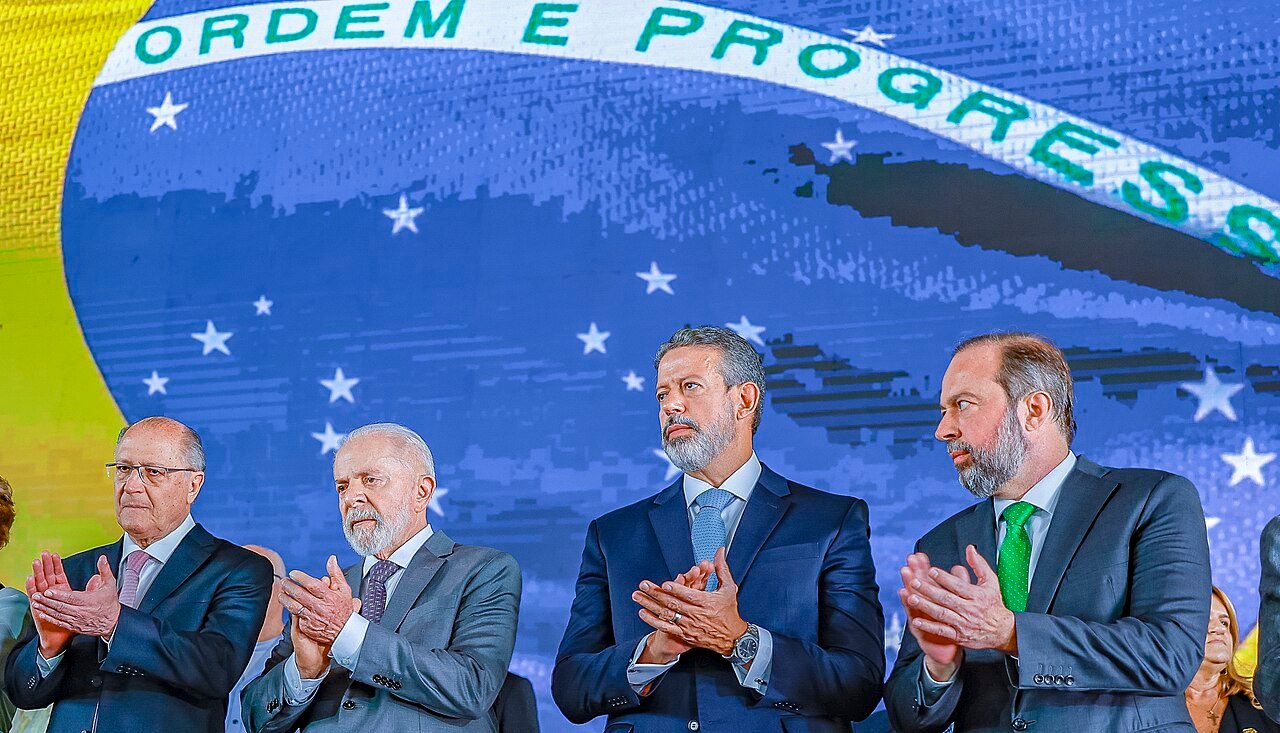 Lula e Geraldo Alckmin aplaudem lado a lado diante de painel com a bandeira do Brasil