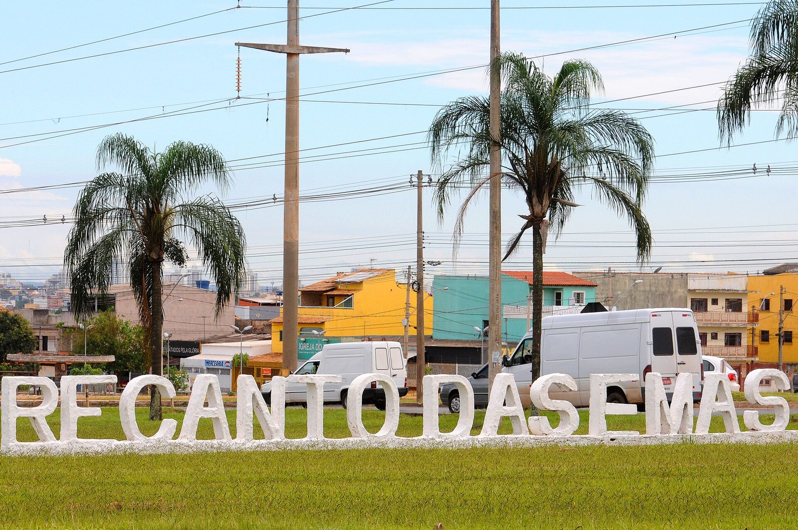 Letreiro de concreto com o nome Recanto das Emas em gramado, com palmeiras e casas ao fundo