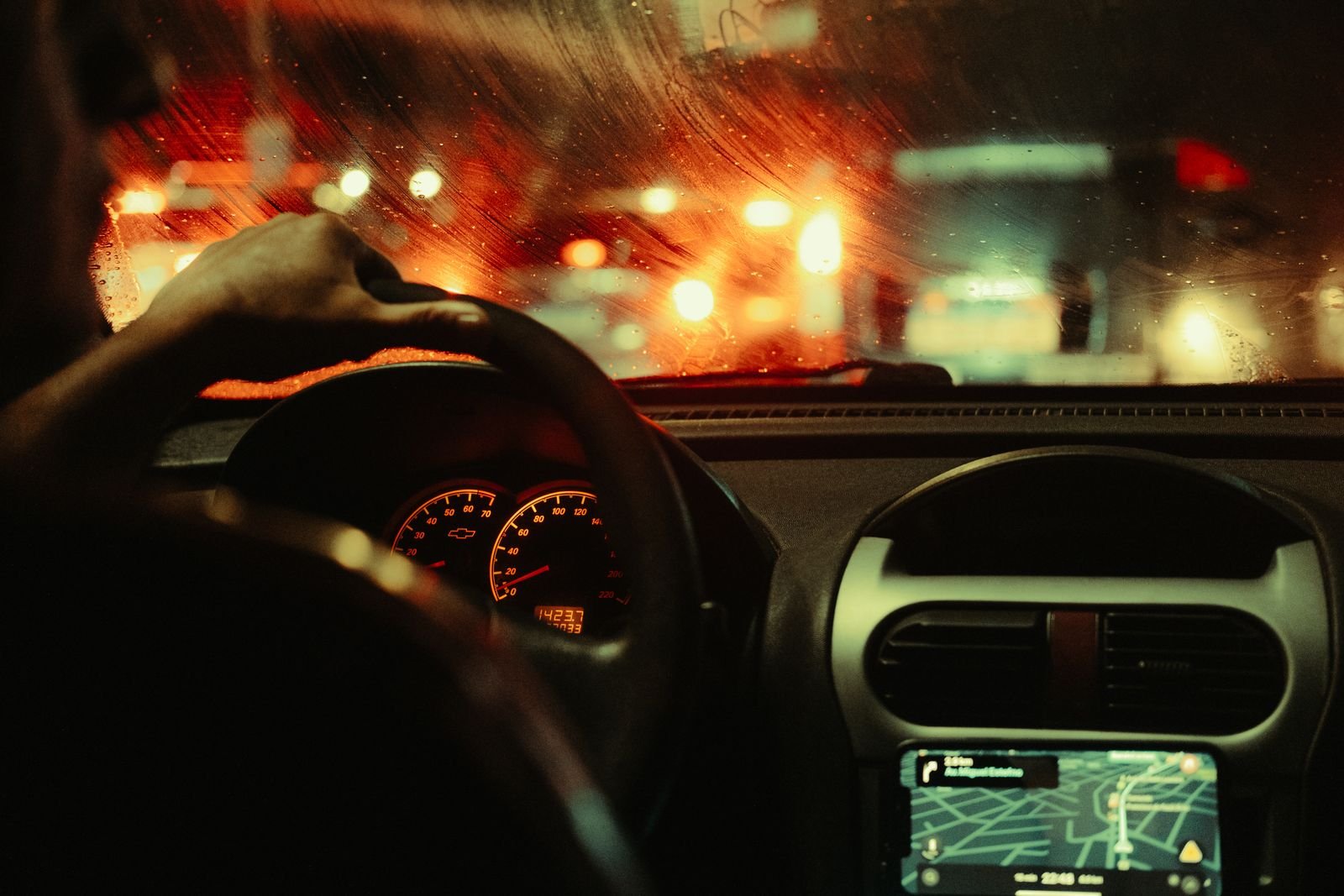 Vista de dentro de um carro à noite, com a mão do motorista no volante e o painel aceso, sob chuva