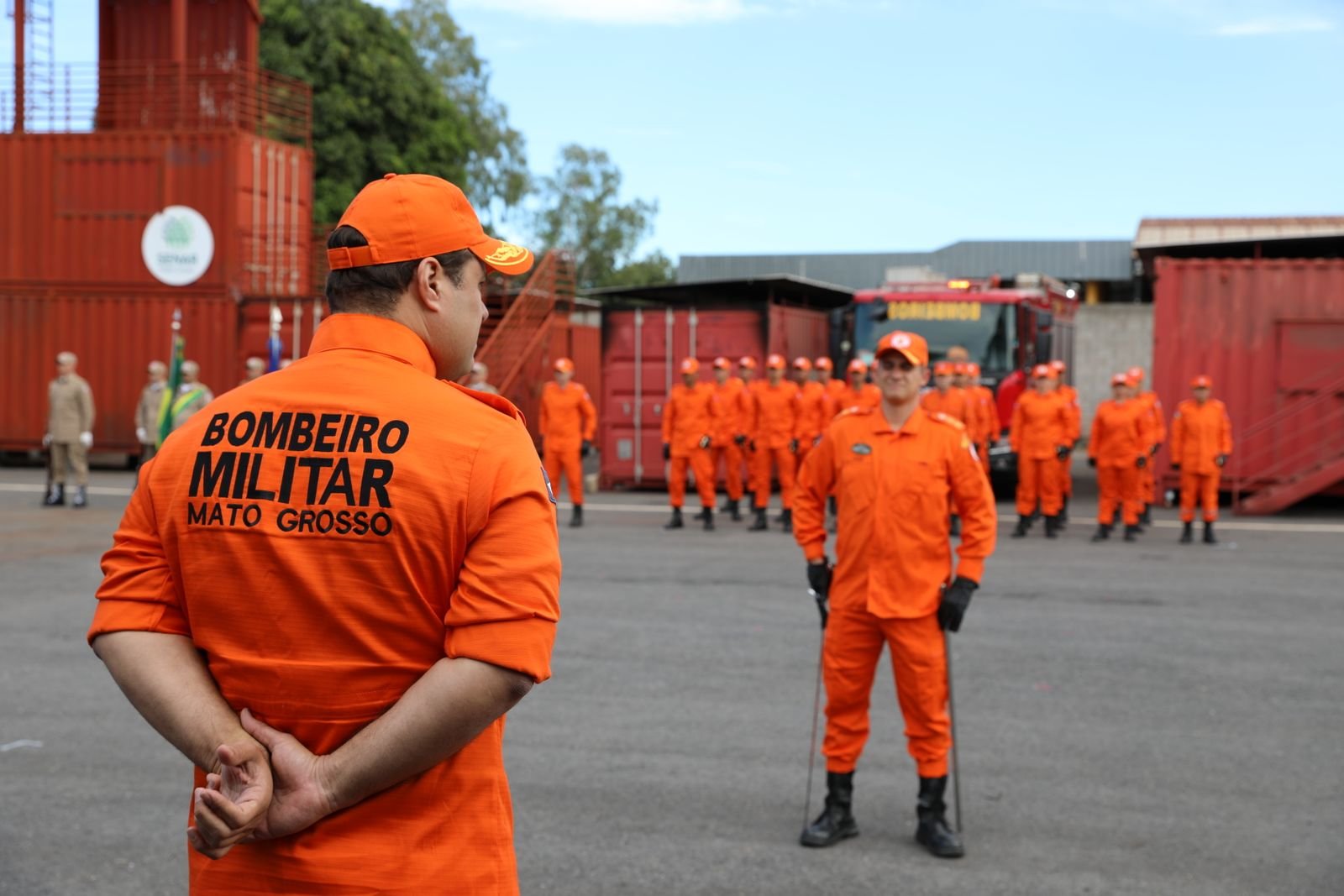 Bombeiros militares de Mato Grosso, de uniforme laranja, em formação diante de contêineres de treinamento e de um caminhão de bombeiros