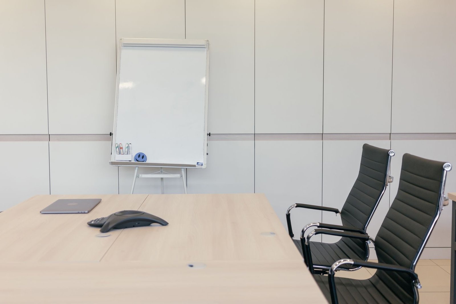 Sala de reunião vazia com mesa de madeira clara, telefone de conferência, cadeiras pretas e quadro branco ao fundo
