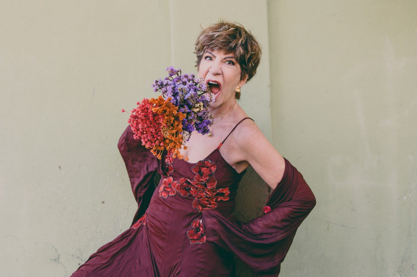 Sandra Pêra sorri com um buquê de flores secas nas mãos, de vestido vinho, diante de uma parede clara