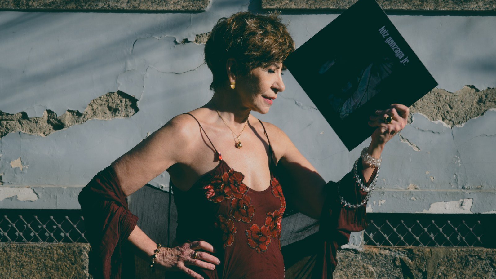 Sandra Pêra, de vestido vermelho, segura e observa a capa de um disco de Luiz Gonzaga Jr. diante de um muro descascado