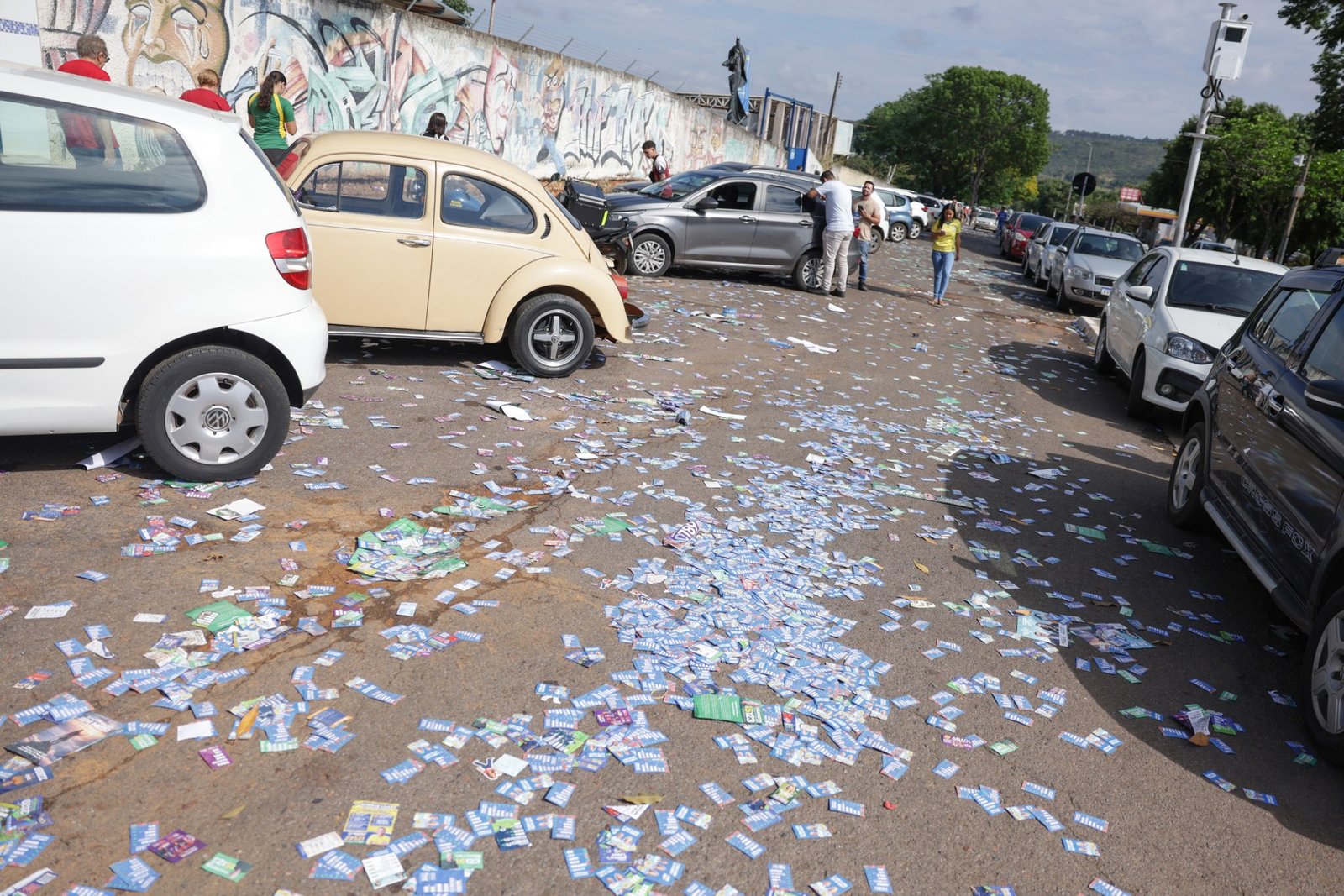 Santinhos de candidatos espalhados pelo asfalto de uma rua com carros estacionados, perto de um local de votação no primeiro turno