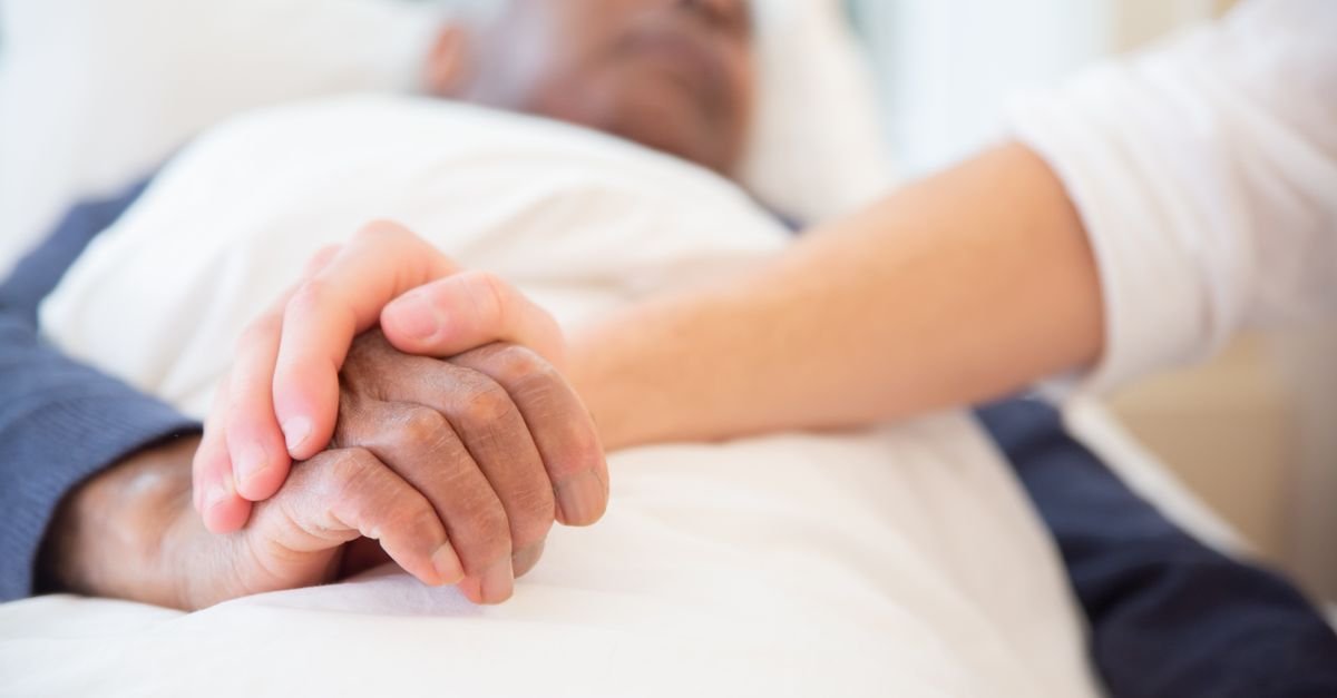 Mãos de uma pessoa segurando a mão de um paciente idoso deitado em leito hospitalar
