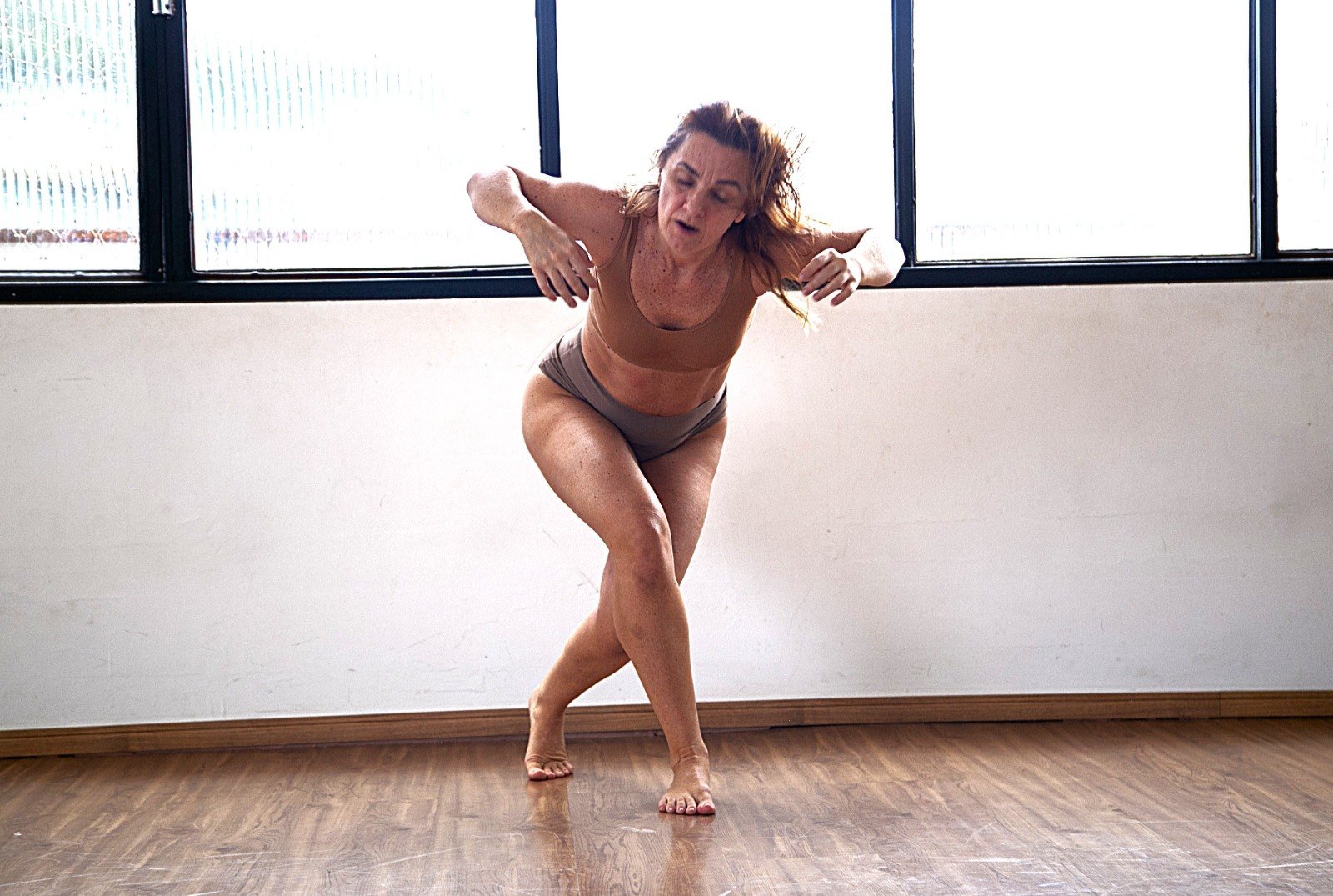 Bailarina em traje de ensaio dança curvada, com os braços abertos, em sala iluminada por janelas