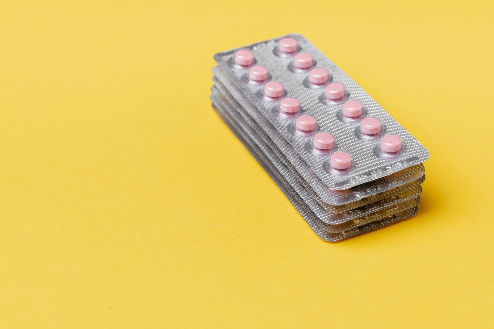 Pilha de cartelas de comprimidos rosa sobre fundo amarelo, imagem ilustrativa de medicamentos
