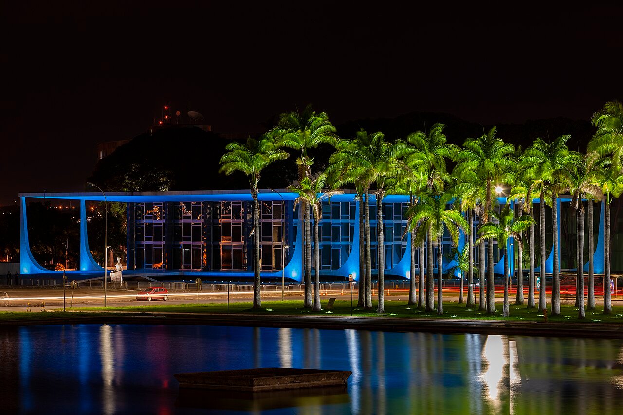 Fachada do edificio do Supremo Tribunal Federal iluminada de azul a noite, com palmeiras e espelho d'agua em primeiro plano