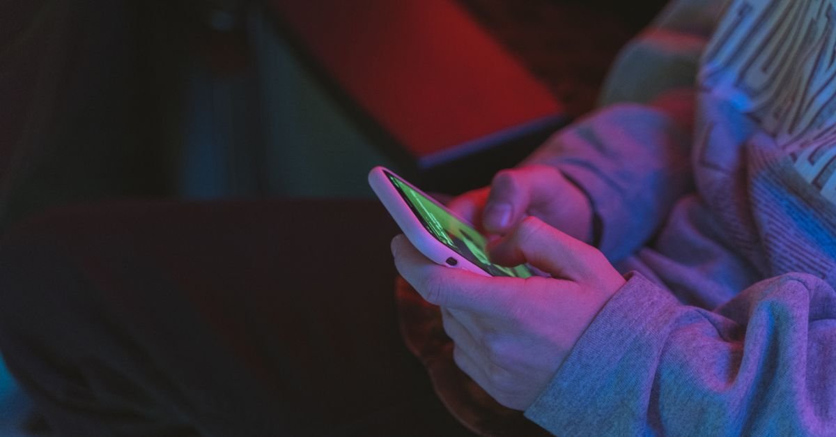 Mãos de uma pessoa jovem segurando um celular com a tela acesa em ambiente escuro
