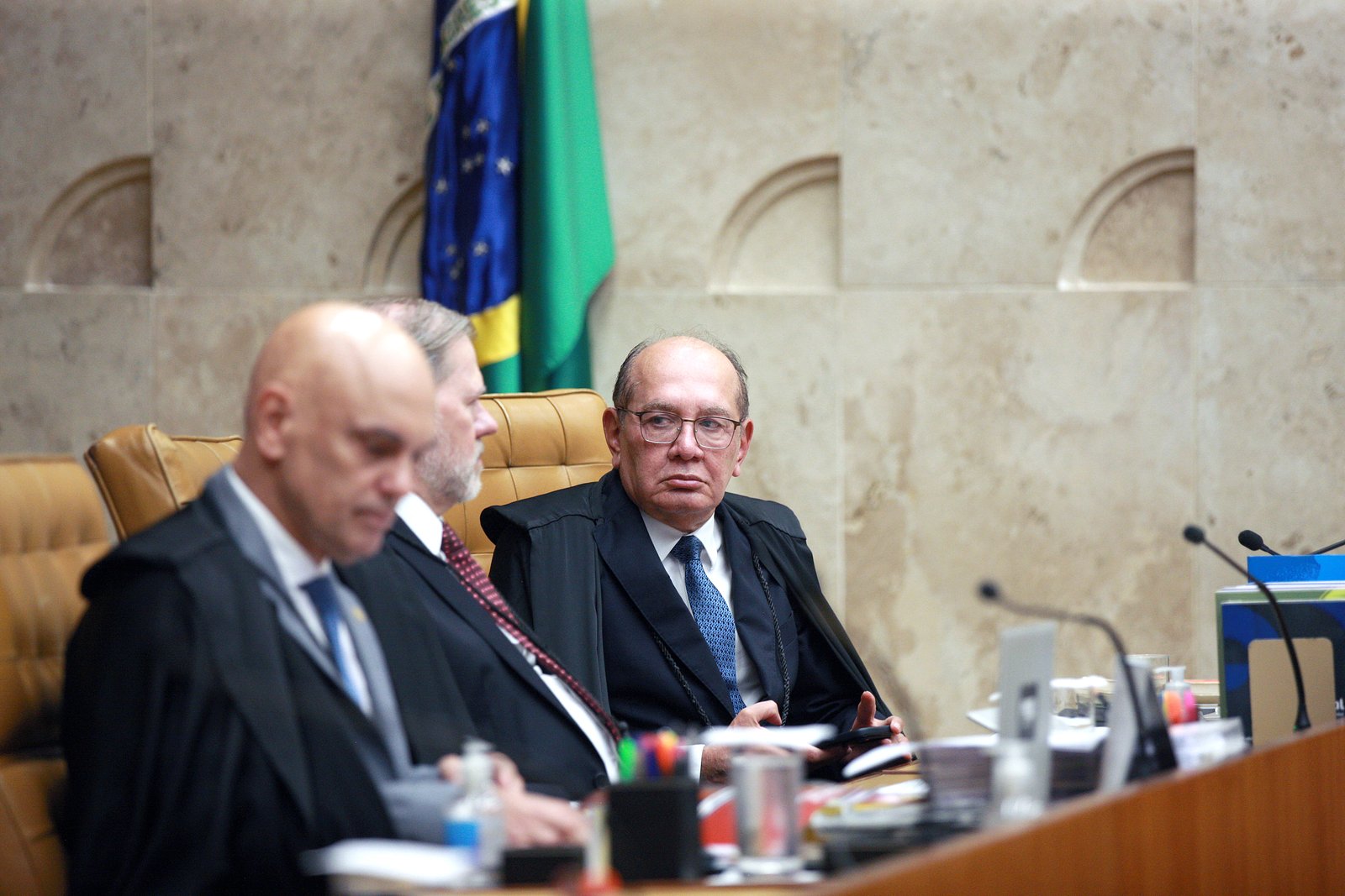 Ministros do Supremo Tribunal Federal sentados à bancada do plenário durante sessão, com bandeiras e a parede de mármore do plenário ao fundo