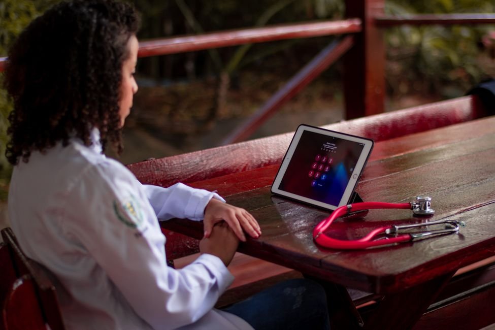 Profissional de saude de jaleco branco sentada a uma mesa de madeira com estetoscopio vermelho e tablet