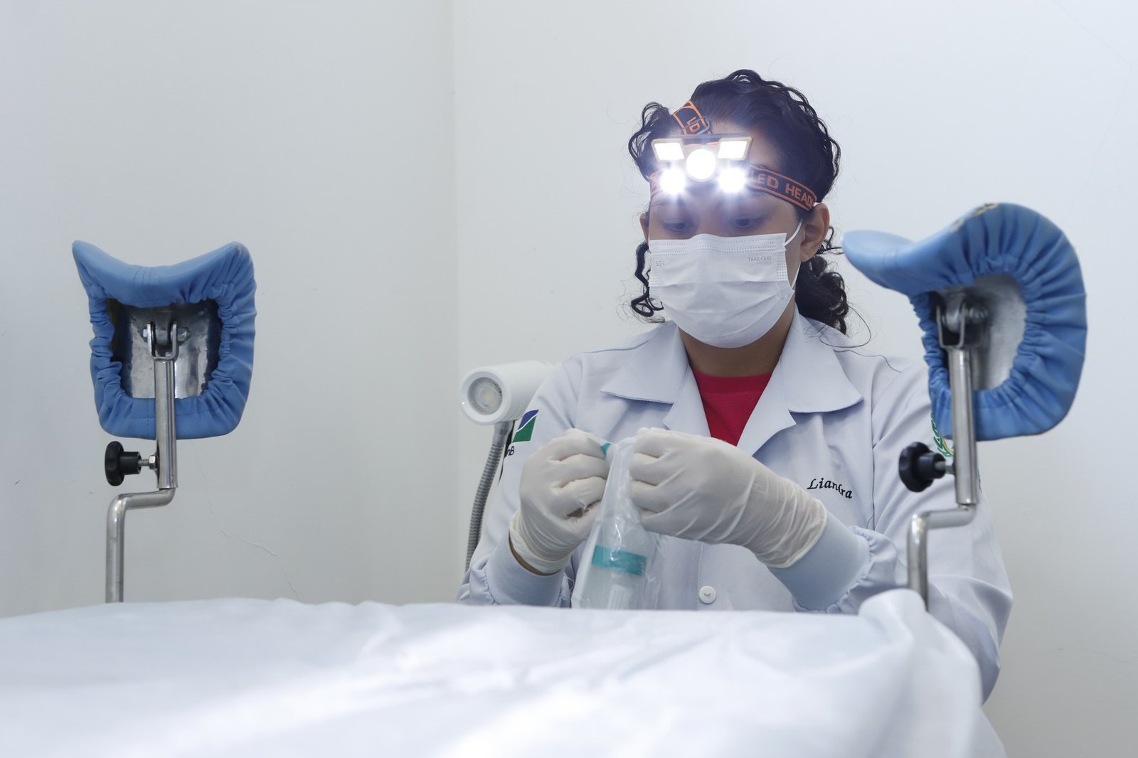 Profissional de saúde com máscara e lanterna de cabeça prepara material de coleta em sala de exame de uma UBS do DF