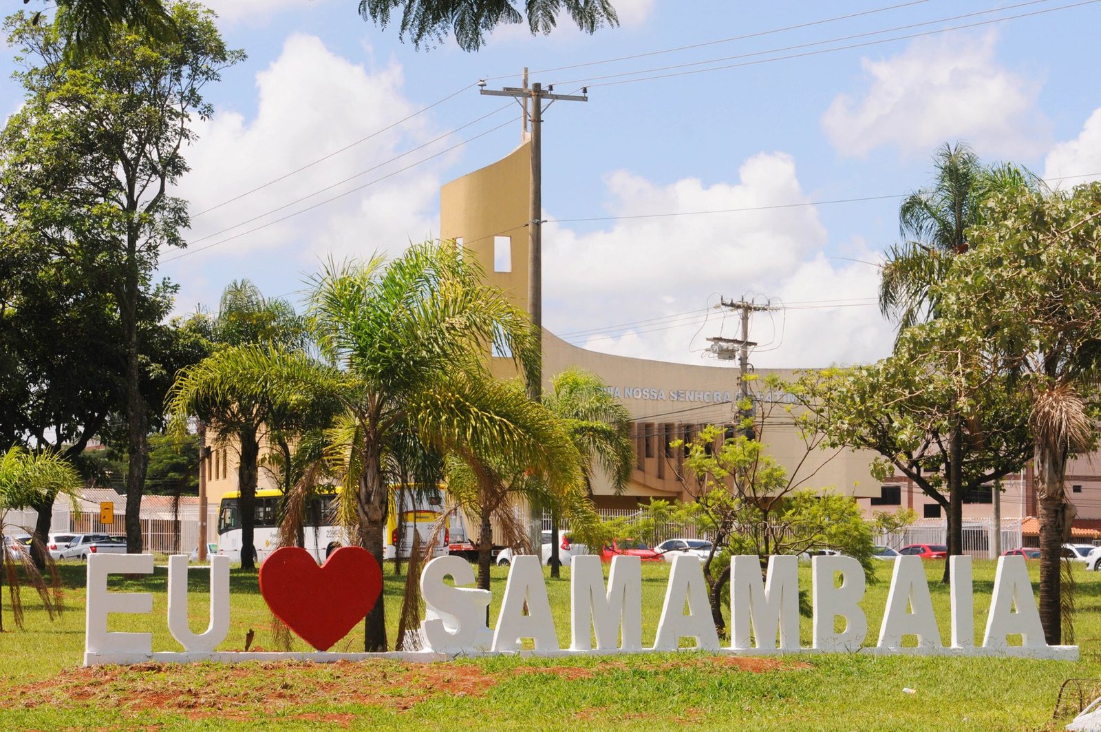 Letreiro com a palavra Samambaia em um gramado, com igreja e ônibus ao fundo