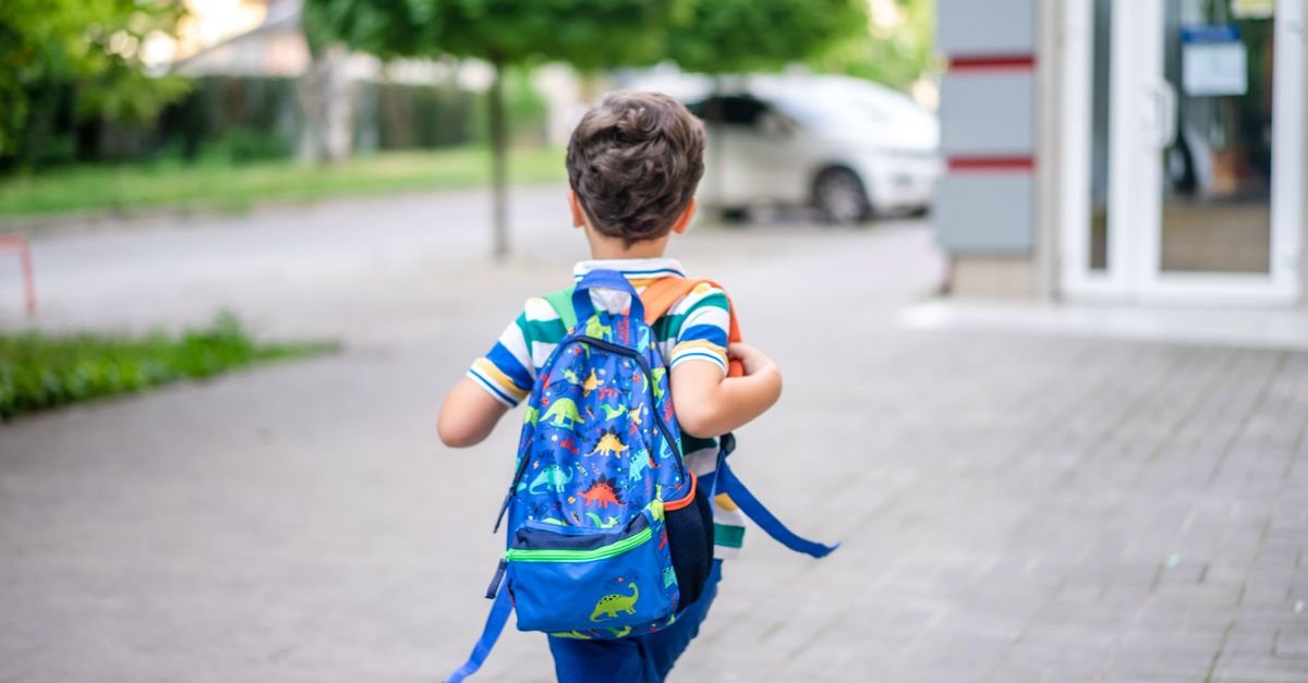 Criança de costas, com mochila de dinossauros, caminha em direção a uma escola, em foto de arquivo
