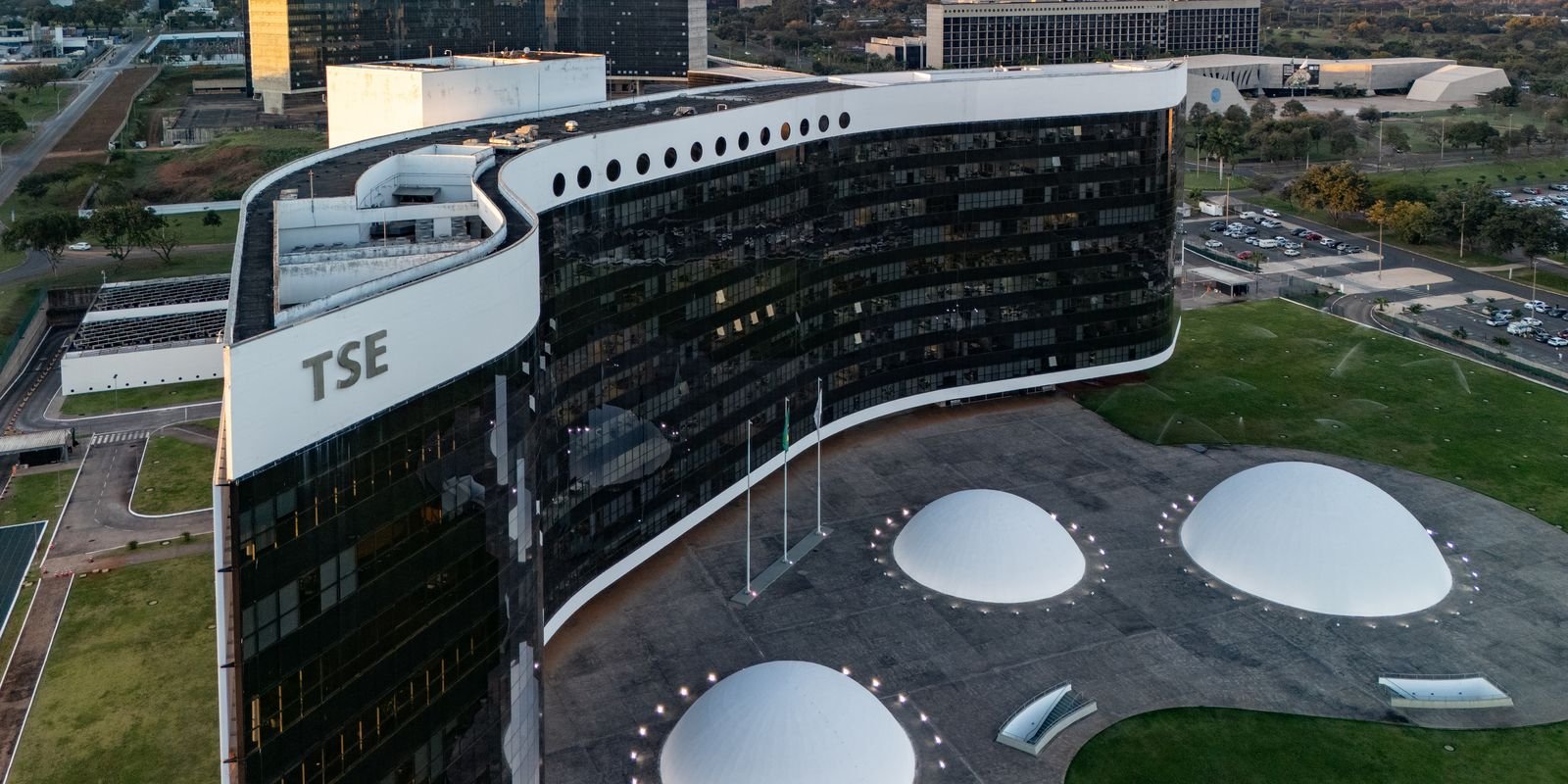 Vista aérea da sede do Tribunal Superior Eleitoral, em Brasília, com a sigla TSE na fachada curva de vidro e as cúpulas brancas da praça