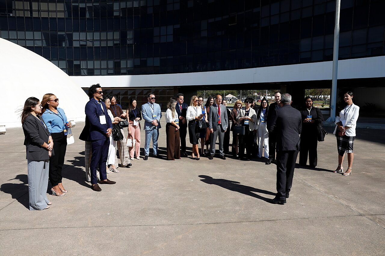 Grupo de visitantes reunido no pátio da sede do Tribunal Superior Eleitoral, em Brasília, com a fachada envidraçada do prédio ao fundo