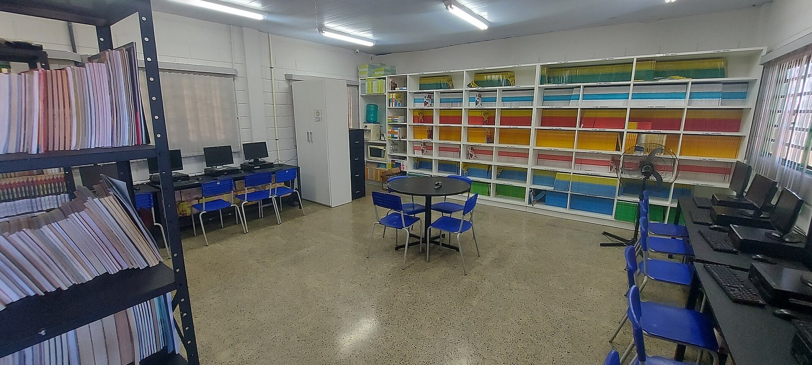 Sala de leitura e informática de escola pública da rede do Distrito Federal, com estantes de livros, computadores e cadeiras azuis