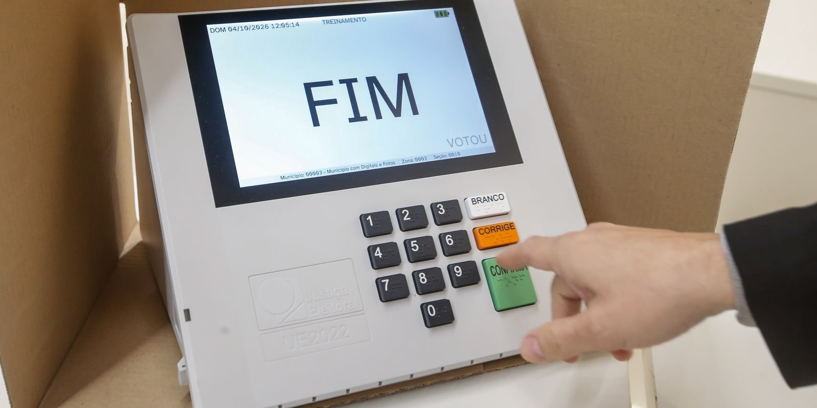 Urna eletrônica em modo de treinamento exibe a tela FIM, enquanto uma mão aponta para o teclado numérico