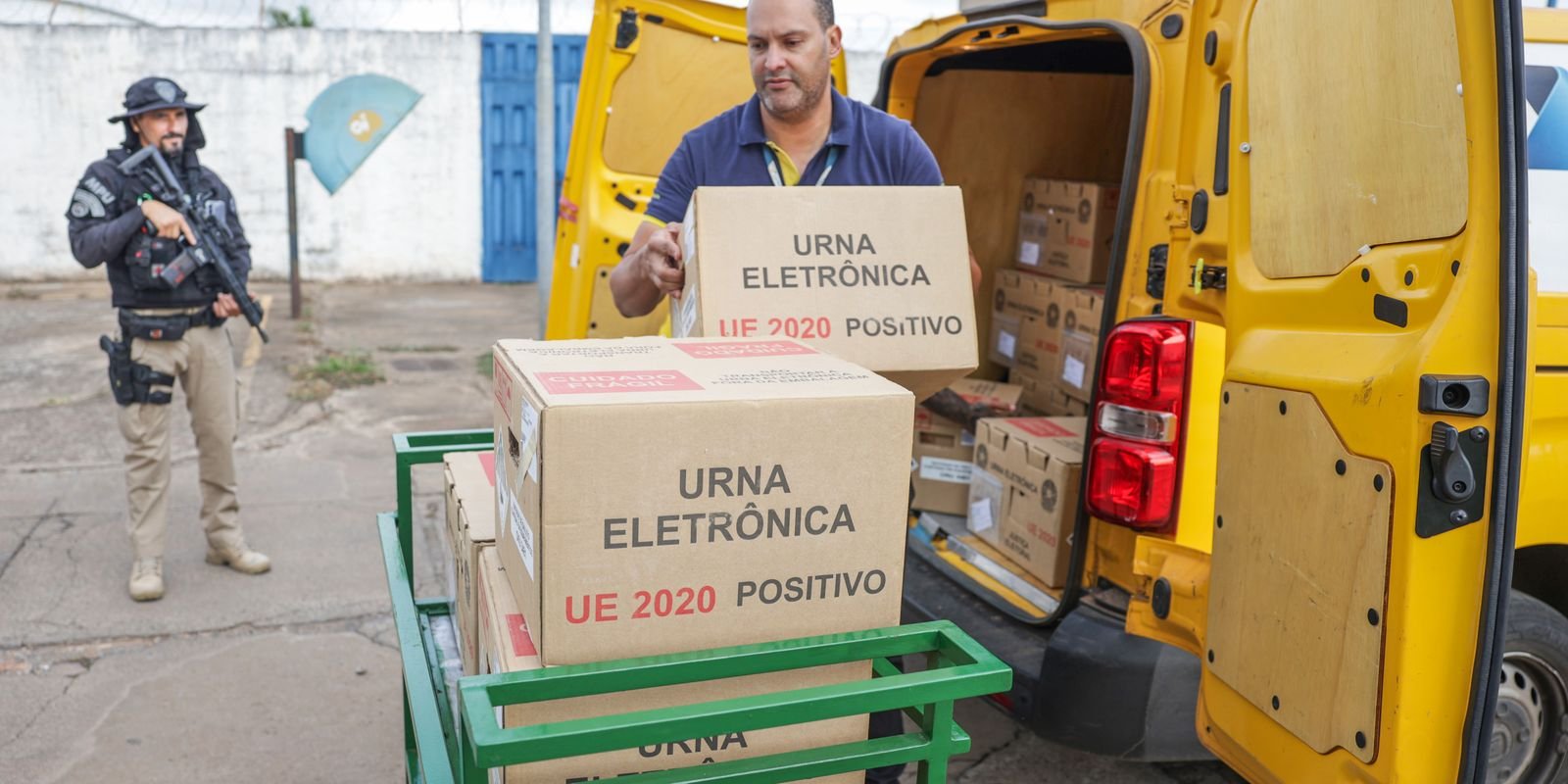 Funcionário da Justiça Eleitoral descarrega caixas de urna eletrônica de uma van amarela, com um agente armado ao fundo