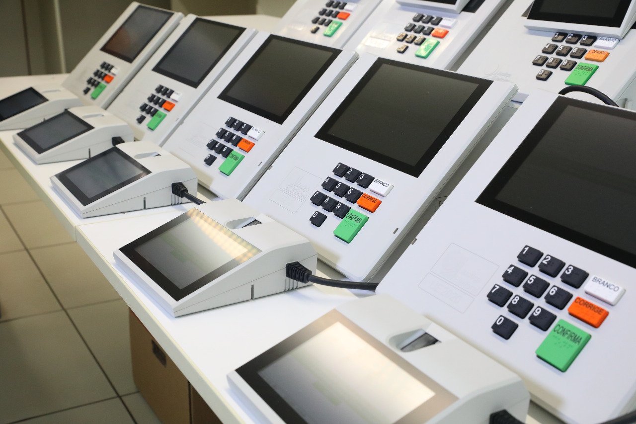 Urnas eletrônicas novas enfileiradas sobre bancada, com teclado numérico e teclas branco, corrige e confirma