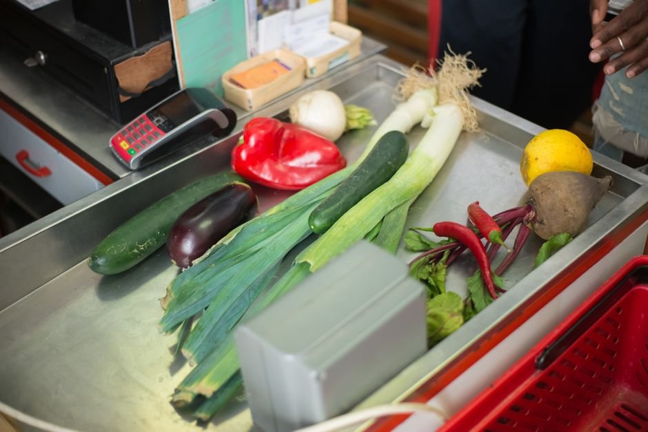 Legumes e verduras sobre o balcão metálico de um caixa de mercado, com cesta vermelha ao lado